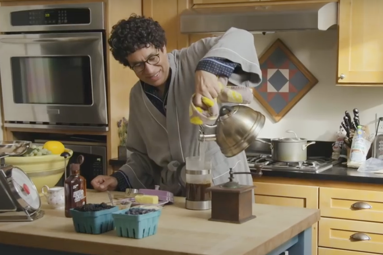 A beaming Fred Armisen, wearing a bathrobe, makes coffee in a rustic kitchen